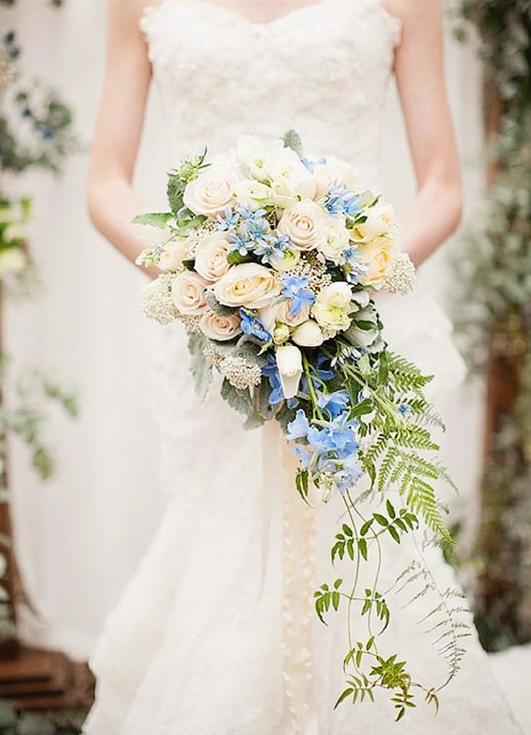 How incredible is this cascading bouquet filled with ivory floral, lush greens, and a magical touch of blue!